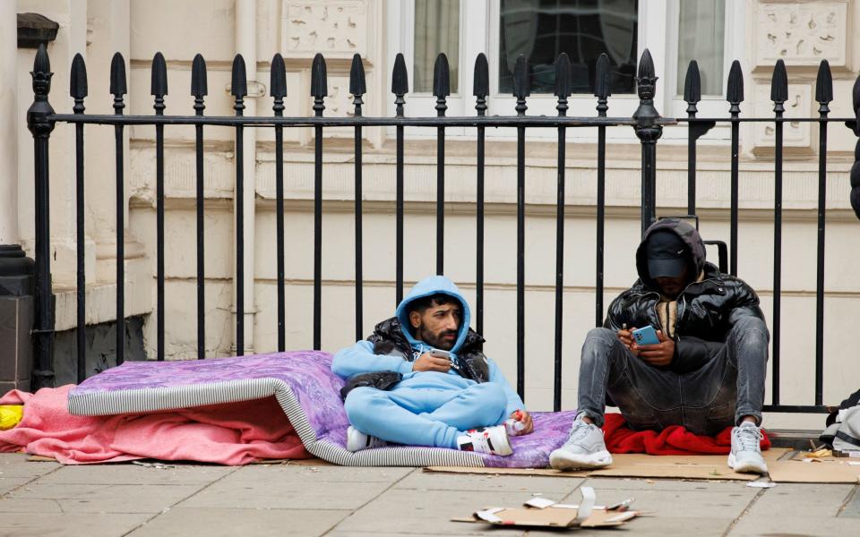 Two men sit and recline on a mattress on the pavement - Jamie Lorriman for The Telegraph