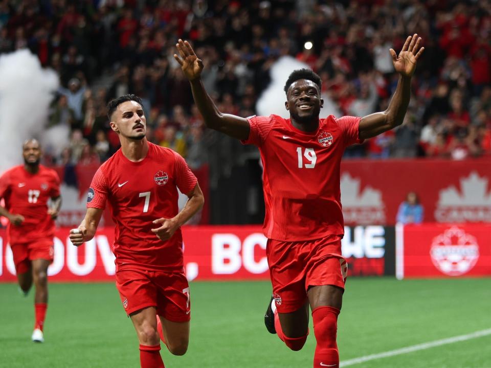 Alphonso Davies raises his hands and smiles while running after scoring a goal as teammates run behind him.