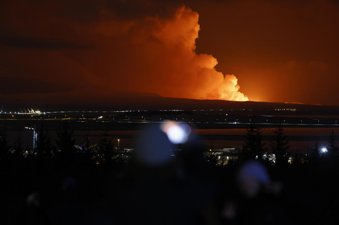 People watch as the night sky is illuminated caused by the eruption of a volcano on the Reykjanes peninsula of south-west Iceland seen from the capital city of Reykjavik, Monday Dec. 18, 2023. (AP Photo/Brynjar Gunnarsson)