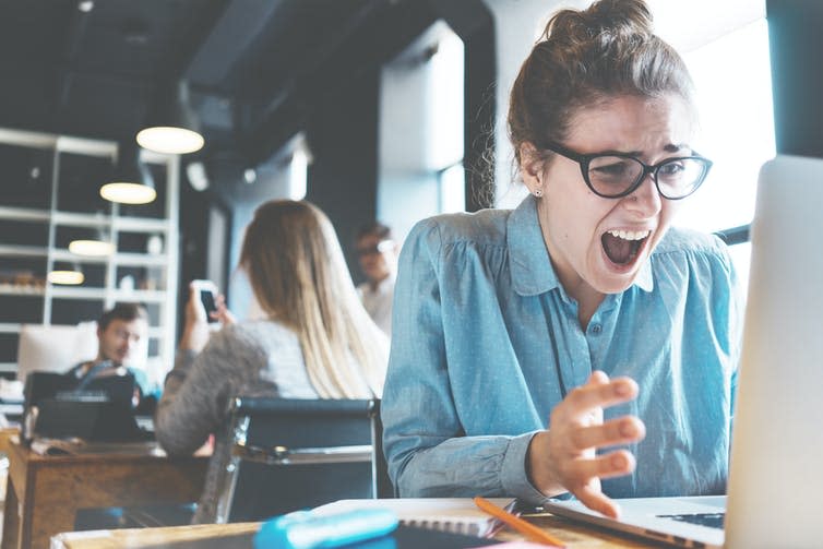 A stressed woman screams at her computer in a busy office.