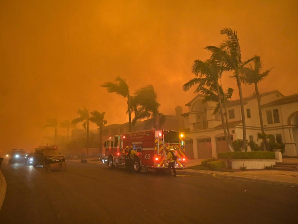 firefighting vehicles drive through orange smoky air on street with homes and palm trees