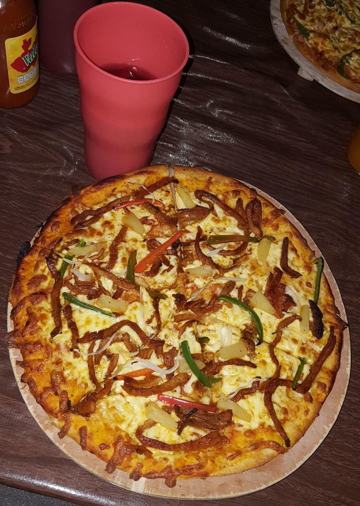 Pizza Al Pastor on a wooden serving board, a soda in a pink plastic cup and a bottle of hot sauce.