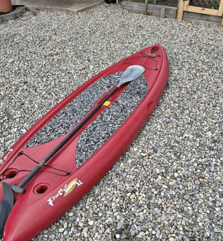 A red kayak named "Freshie" on a bed of small gray pebbles, with a black and gray paddle resting on top