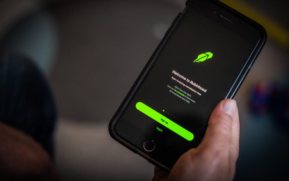 A smartphone shows the black and lime green introduction screen of the Robinhood app, held in a calloused pink hand in front of a blurred background - Tiffany Hagler-Geard/Bloomberg
