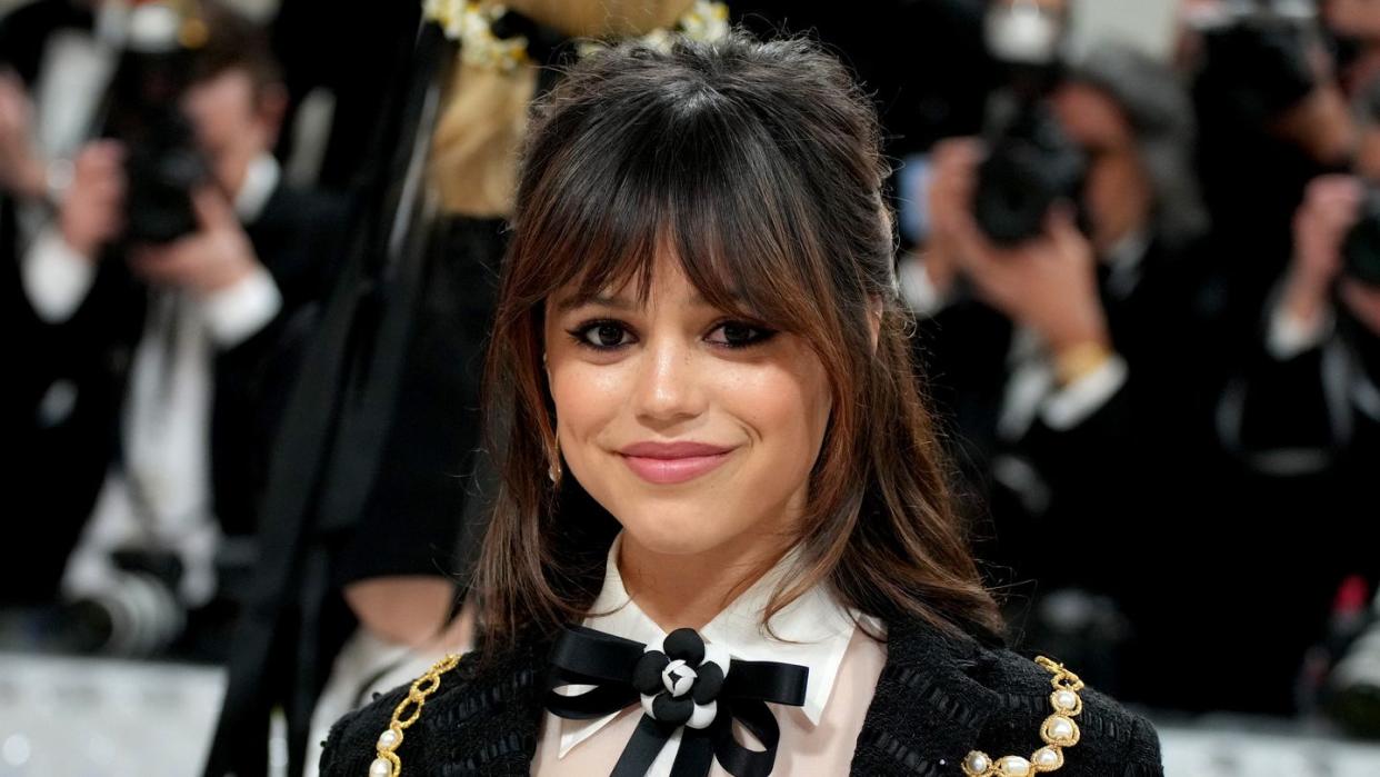 jenna ortega smiles at the camera, she wears a black and white formal outfit with a black bow in her white collared shirt, she has shoulder length brown hair and bangs