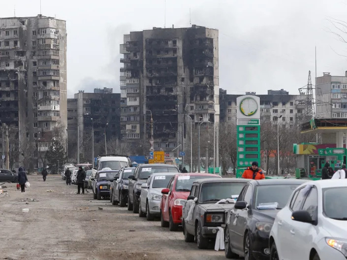 cars of evacuees line a road leaving blocks of burnt out destroyed apartment buildings