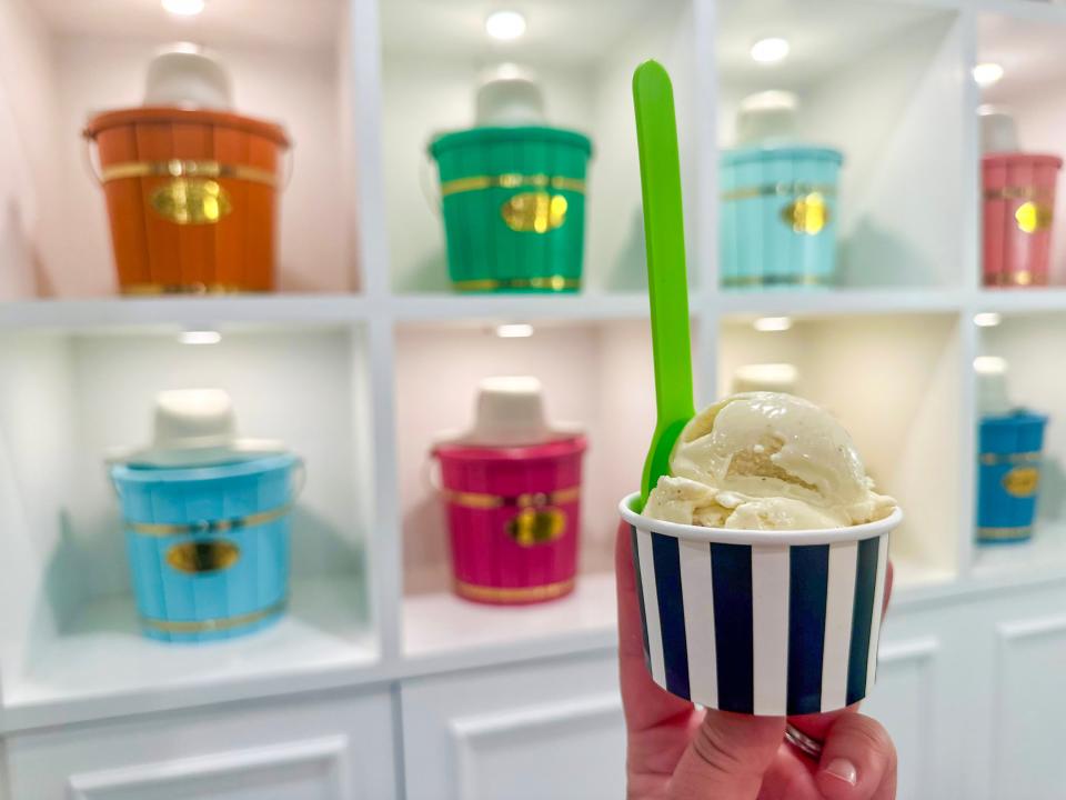 A hand holds a small black and white striped cup of vanilla ice cream with a green spoon sticking out in front of several colorful buckets on white shelves at ice-cream shop