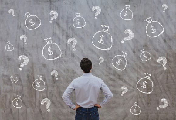 A man staring at a wall covered with drawings of bags of money and question marks.