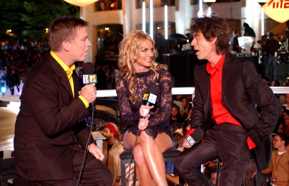 A man with a microphone talks to another man and a smiling blond woman looks on.