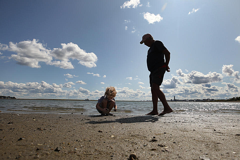 A man stands on a beach next to a child, squatting and apparently inspecting the sand.
