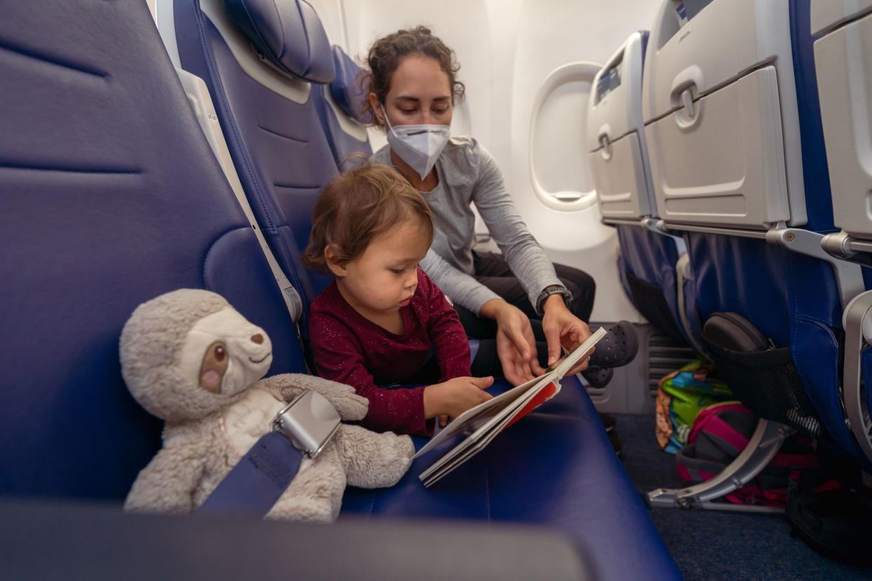 A young mom of Pacific Islander descent wearing a protective face mask reads a book to her Eurasian toddler daughter while riding on an airplane. The woman is wearing a protective face mask.