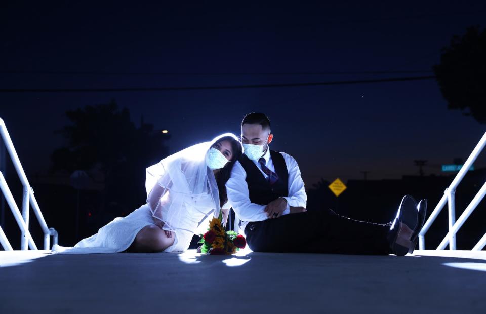 A man in vest and tie and a woman in a wedding dress and veil sit together, wearing masks.