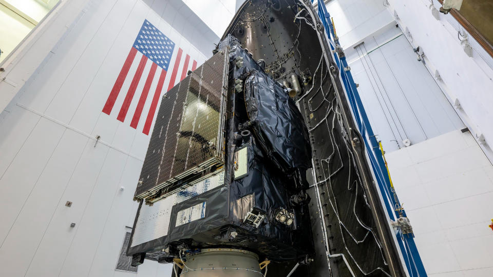 a satellite is seen before being encapsulated inside its rocket's payload fairing, inside a large, white-walled assembly building.