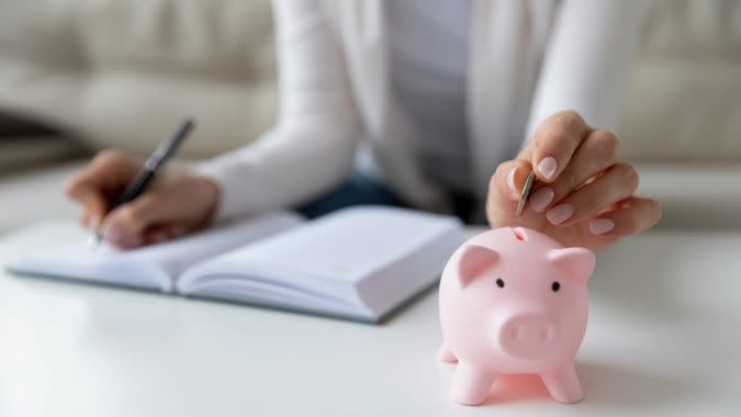 Thrifty woman sit at table hold pen writing daily expenses in diary put coin in pink piggy bank close up.