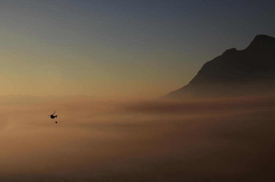 A helicopter hovers above the smoke which engulfs the city of Cape Town, South Africa, Tuesday April 20, 2021. A massive fire spreading on the slopes of the city's famed Table Mountain, at right, is kept under control as firemen and helicopters take advantage of the low winds to contain the blaze. (AP Photo/Nardus Engelbrecht)