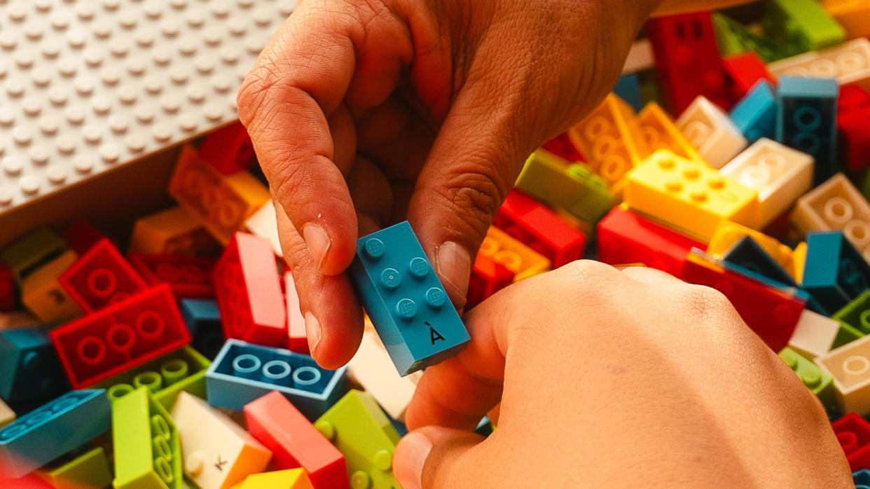  An open box of LEGO® Braille Bricks. A hand holds up a blue LEGO Braille Brick. Another hand is poised to pick a LEGO Braille Brick from the box. 