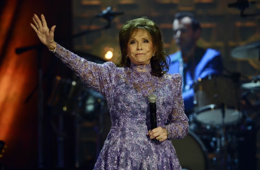 A woman with red hair holding a microphone and waving from a stage in a long-sleeve floral gown