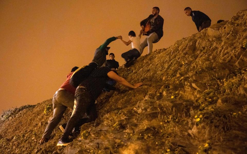 Moroccan migrants climb rocks on the shoreline in the northern town of Fnideq, with a policeman watching from the top of the slope -  FADEL SENNA/AFP via Getty Images