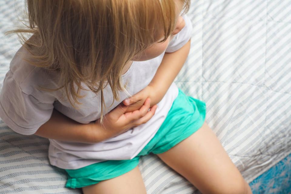 Child sits on the end of a bed, clutching their stomach.