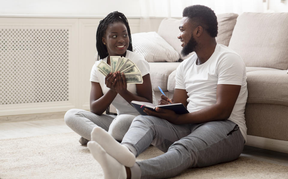 Planning family budget. Happy afro couple sitting on floor at home with dollar cash and notepad, deciding together how to spend money