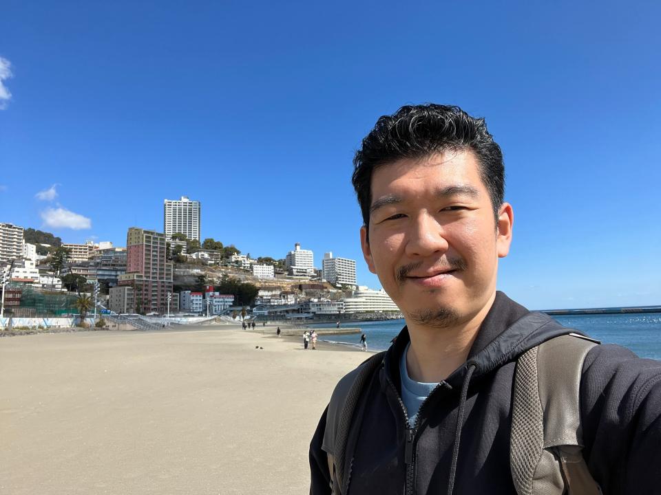 A man takes a selfie with the beach of Atami in the background.