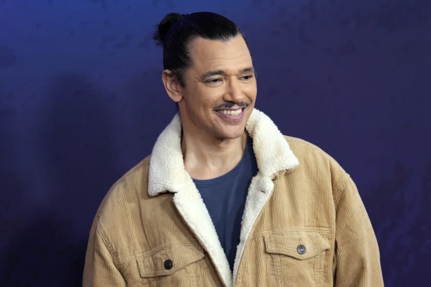 A man with black hair pulled up in a bun and a mustache smiling in a beige jacket and blue shirt
