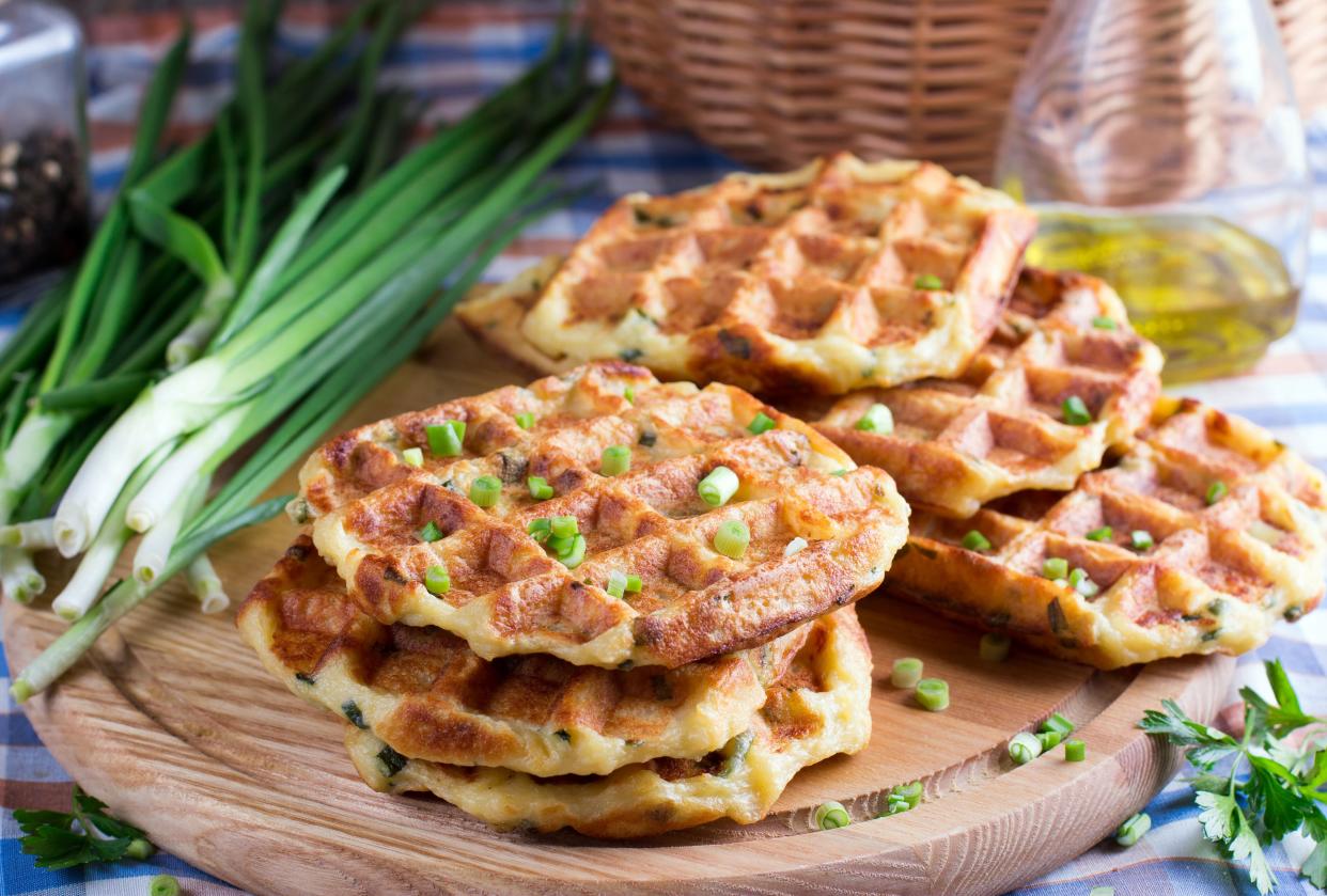 Six loaded potato waffles with chives on a round cutting board with ingredients in the background