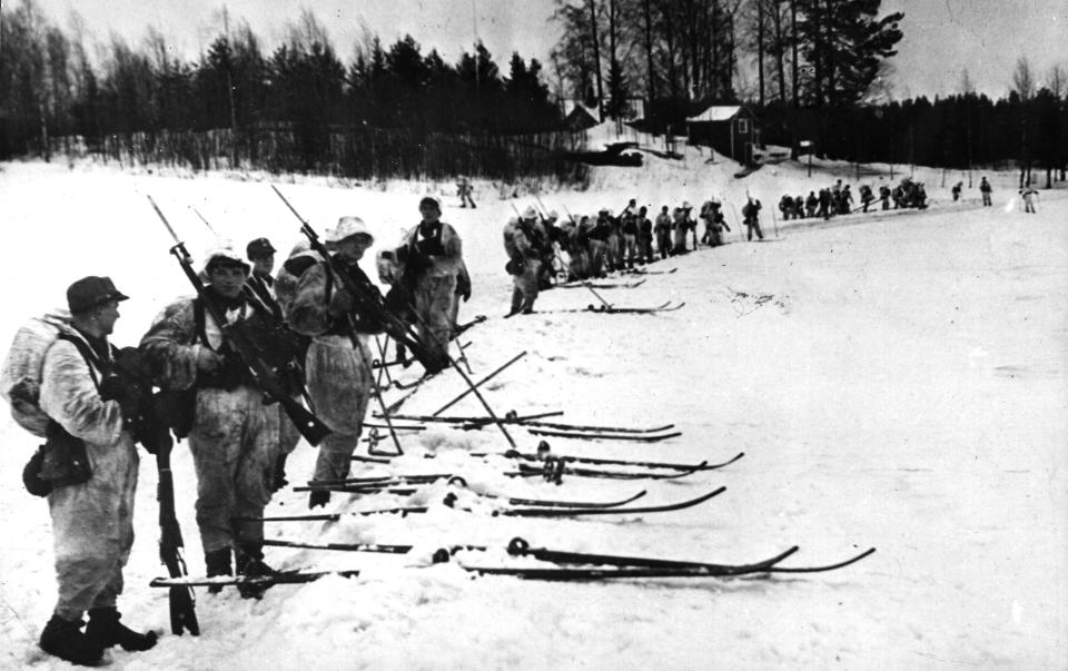 Black-and-white image of several dozen people in military gear with rifles on skis standing in a row.