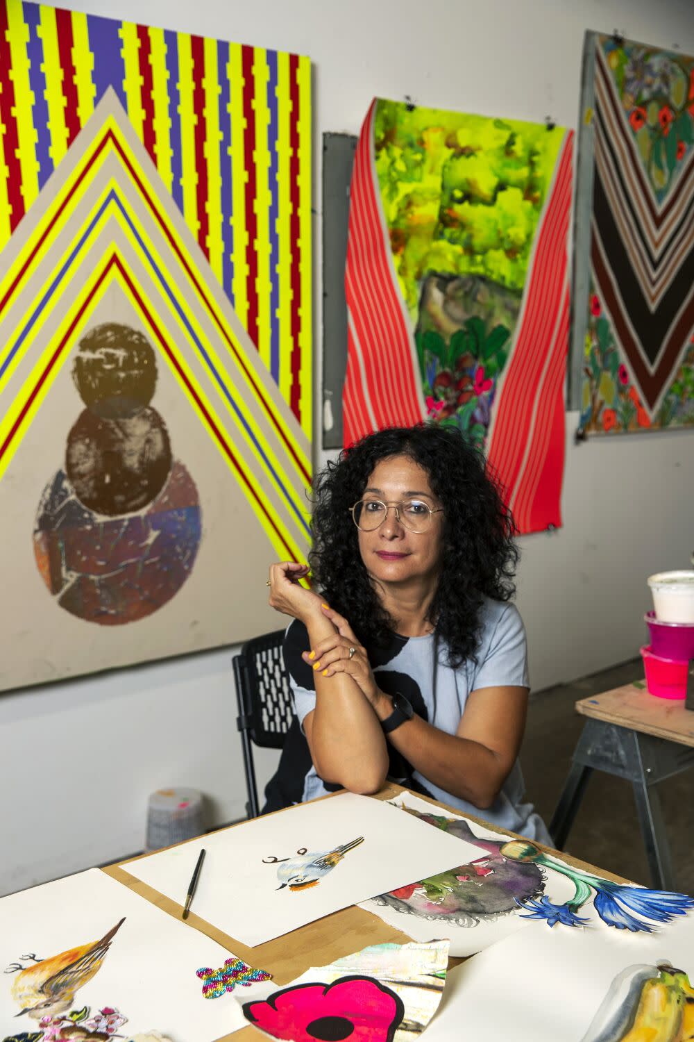 Carolyn Castaño is seen seated in her studio, surrounded by paintings bearing bright patterns and landscape elements