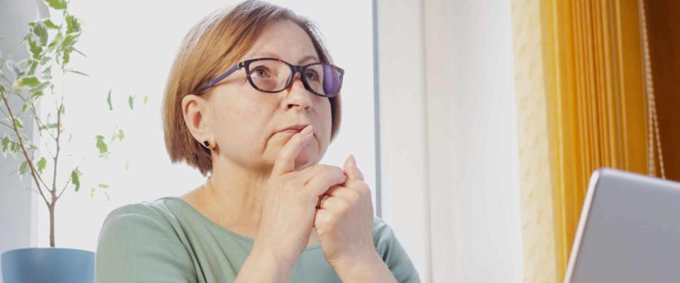 Thoughtful mature woman in eyeglasses with laptop, looking in distance.