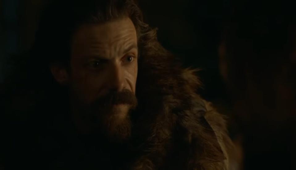 A bearded man in a fur coat stares intensely in torchlight