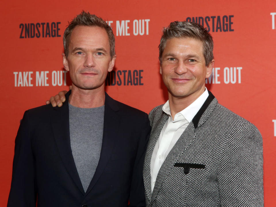 Two men standing close, one in a grey blazer and white shirt, the other in a casual sweater and shirt