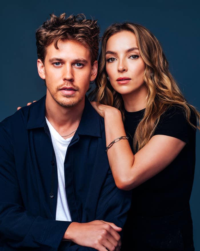 Austin Butler and Jodie Comer, both in casual outfits, pose closely against a blue background. Jodie has her hand on Austin's shoulder