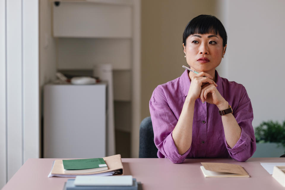 A pensive Asian businesswoman sitting at her desk with a thoughtful expression, illustrating a story on mid-career switch..