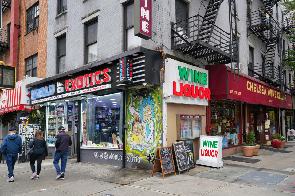 Exterior of "World Exotics" next to "Chelsea Wine Cellar" and "Wine Liquor" shops on a city street. Three people are walking past the stores