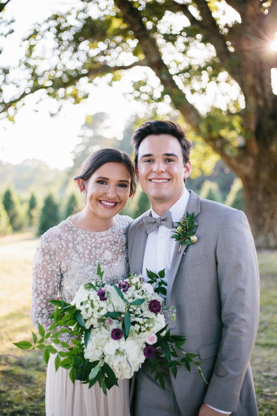 A bride and groom lean together and grin in front of a tree.