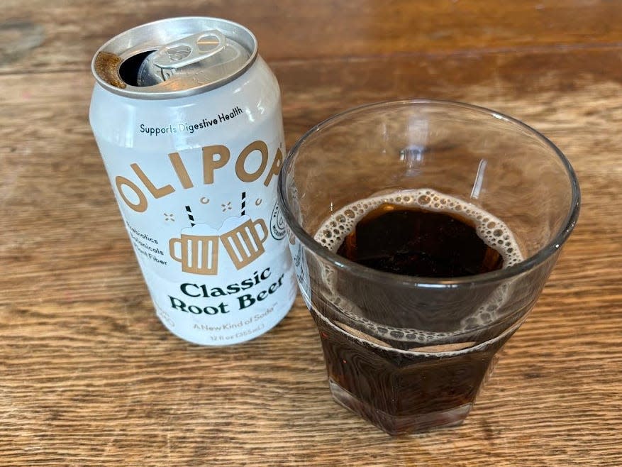 An open can of classic root-beer Olipop next to a small, clear glass with brown liquid inside. Both are sitting on a wooden table.