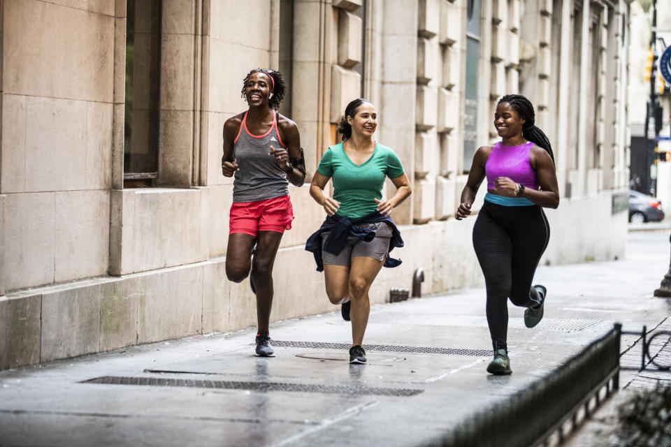 Three women running together down a city sidewalk, smiling and wearing athletic clothing