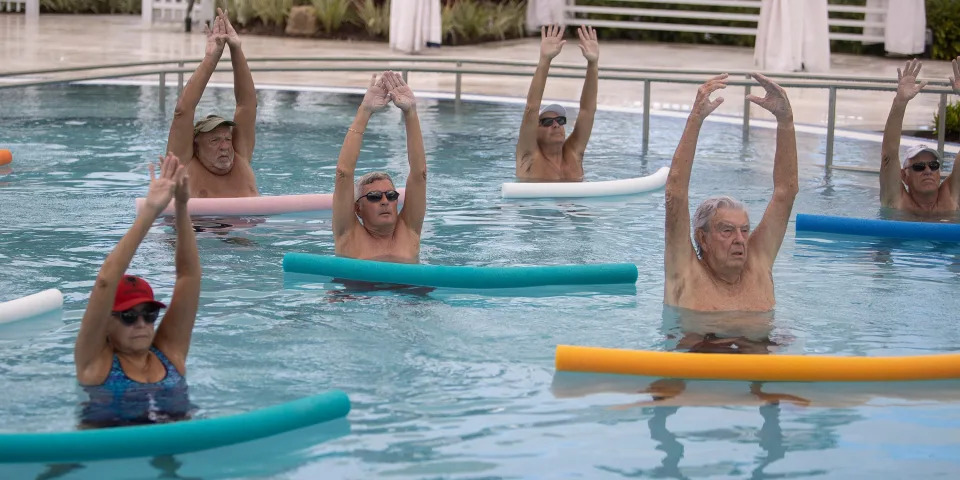 Six retired people are in a swimming pool with their arms over their heads. They each have a pool noodle in front of them.