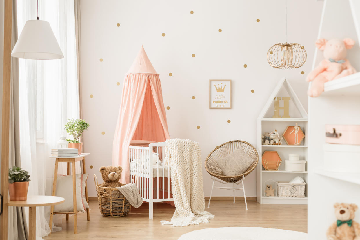 Canopied cradle between gold armchair and basket with teddy bear in pink baby's bedroom interior. (Photo: Getty)