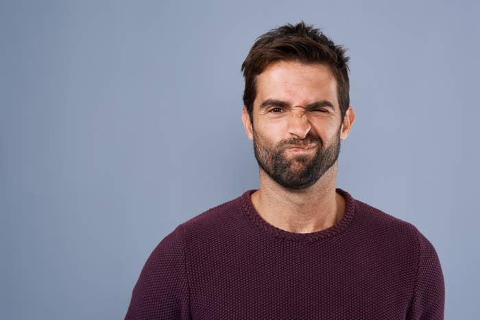 A bearded man squints one eye and makes a funny, scrunched face. He is wearing a textured crewneck sweater