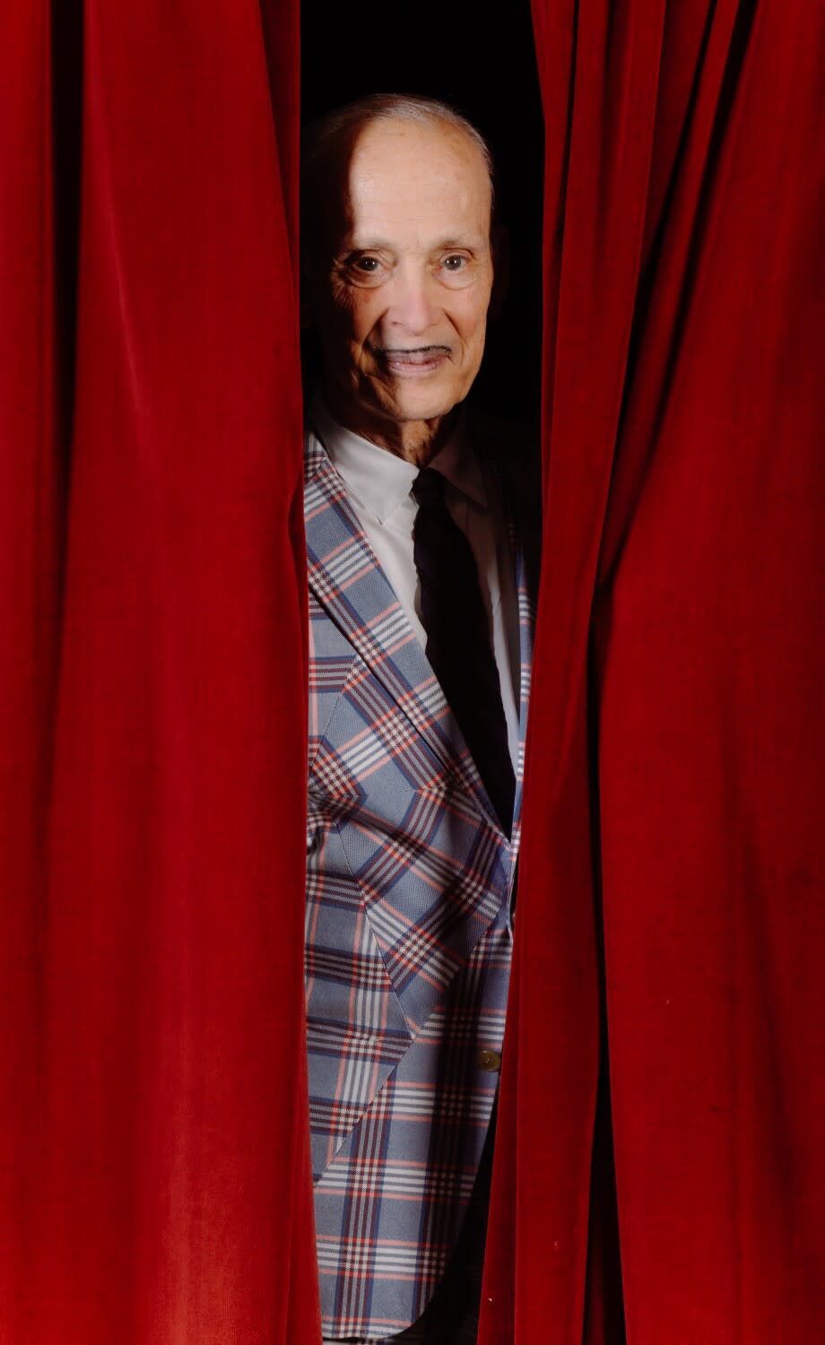 A man in a plaid suit peeks through red curtains.
