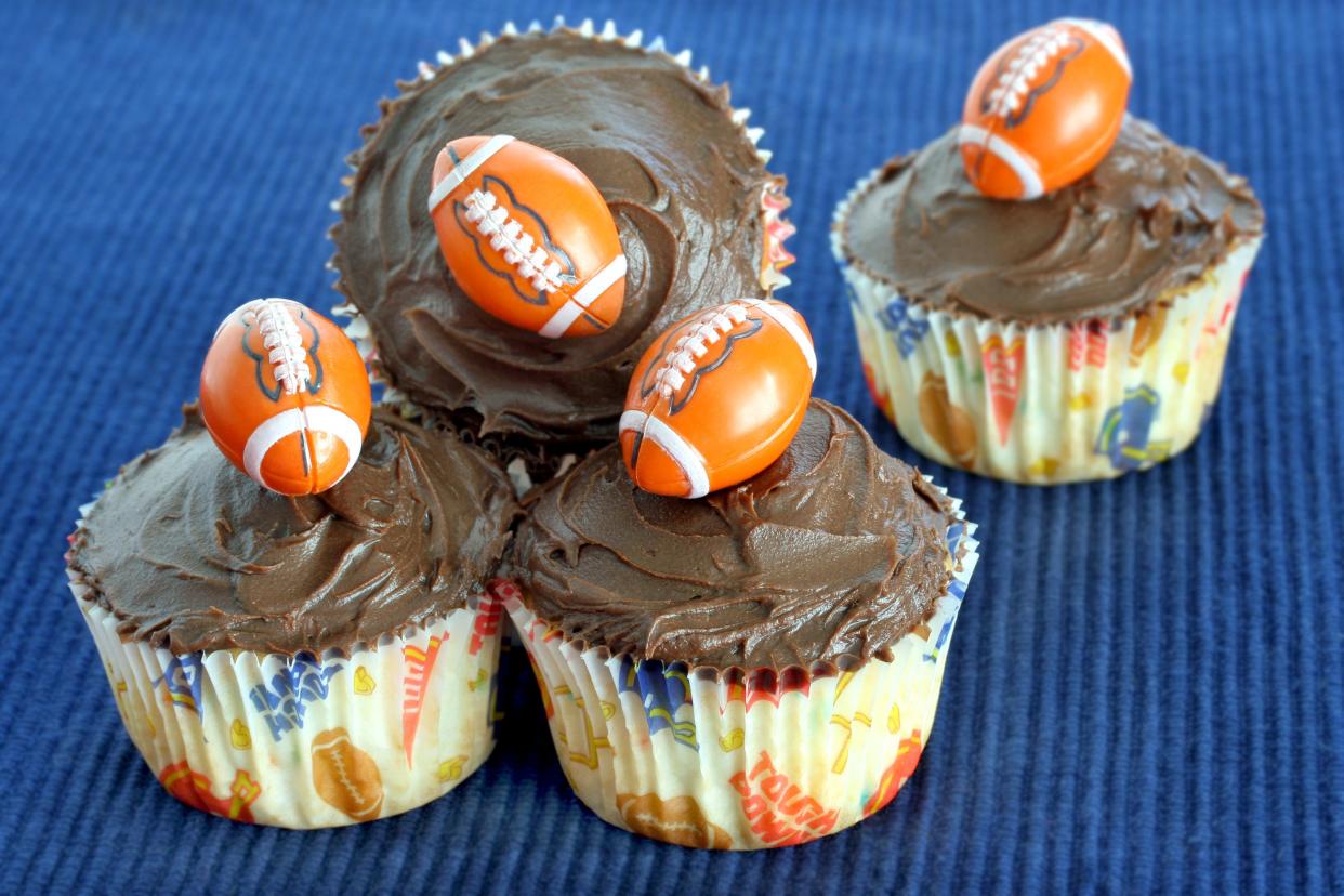 Four vanilla cupcakes with chocolate frosting and mini footballs and football themed liners on a blue background