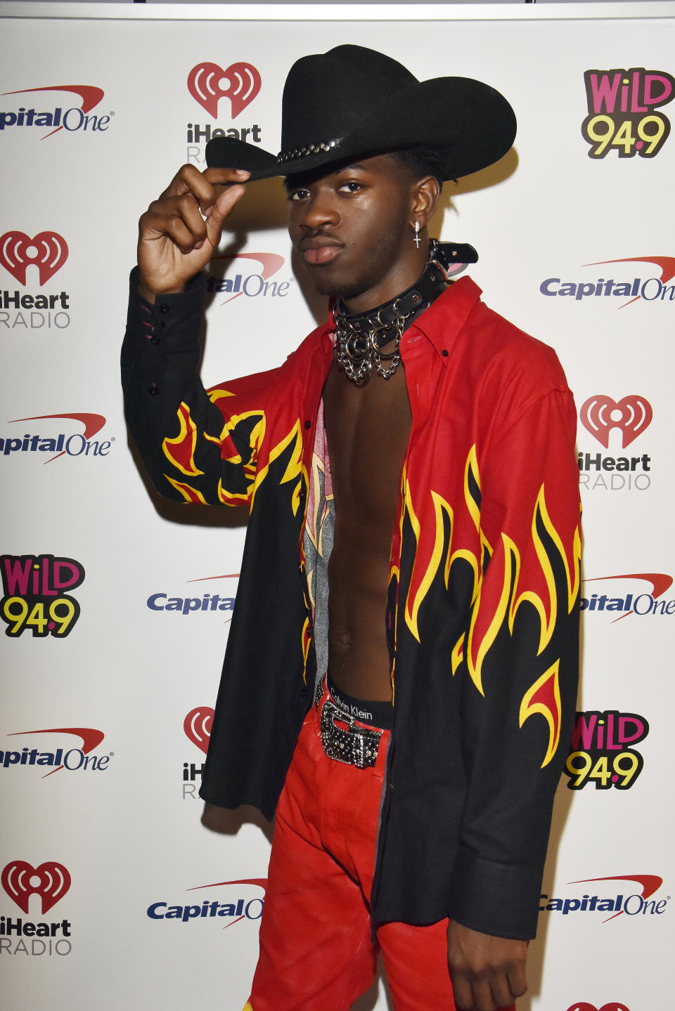 Lil Nas X poses in a black cowboy hat, open flame-patterned shirt, and red pants at an iHeartRadio event