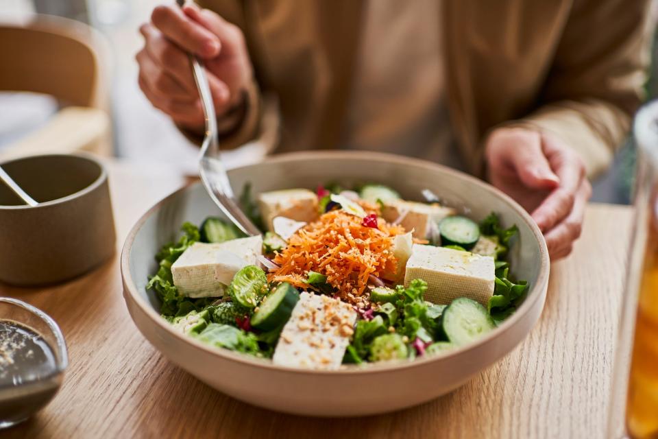 A close-up of a bowl of tofu with veggies — in the background, a woman is sitting at the table to have lunch