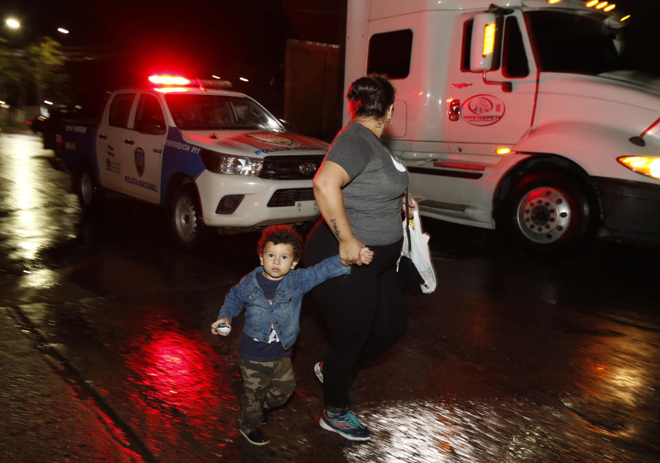 Una mujer con un niño que forma parte de una caravana de migrantes que buscan llegar a Estados Unidos camina frente a un auto policial desde la central de autobuses de San Pedro Sula, Honduras, la noche del lunes 14 de enero de 2019. (AP Foto/Delmer Martínez)