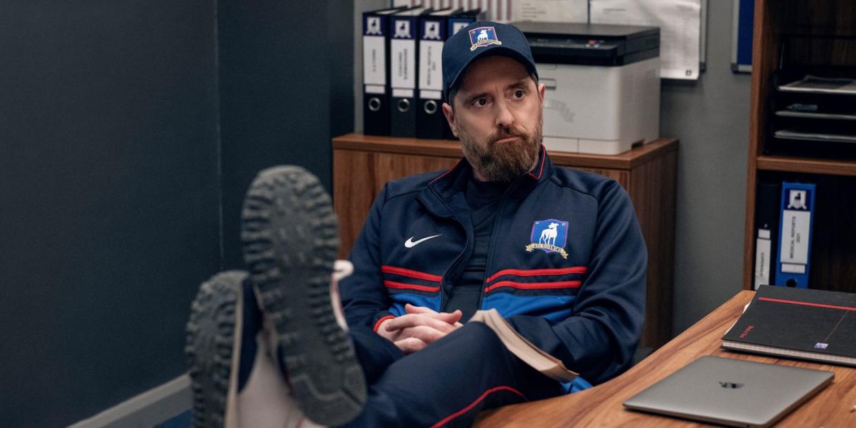 in a scene from ted lasso season 3, coach beard sits with his feet up on the desk in the team office