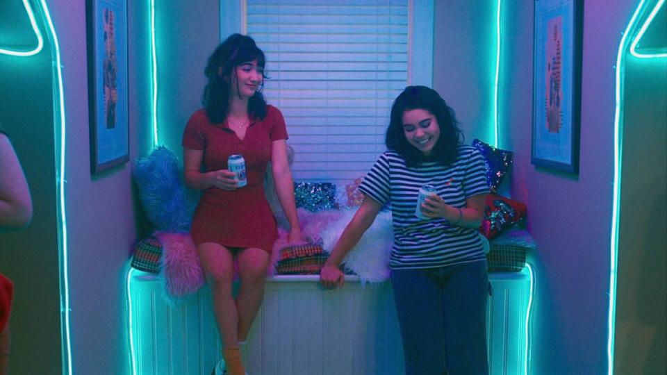 Two women, one in a red dress and one in a striped shirt, sit and chat in a cozy, mood-lit room. One woman holds a beverage can; the other, a smartphone