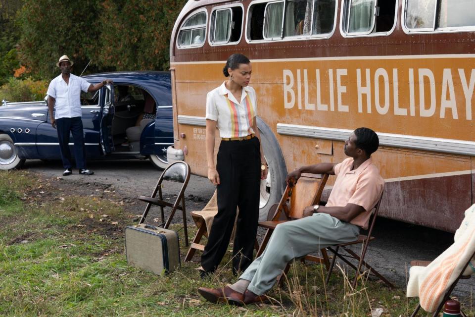 Andra Day as Billie Holiday and Trevante Rhodes as Jimmy Fletcher in a movie scene in the grass near a parked bus.