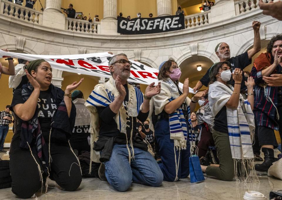 Protesters wearing tallits kneel in a rotunda. A sign reading ceasefire hangs above them against railing.